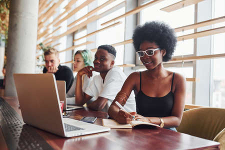 Black woman sitting in front of group of multi ethnic people with alternative girl with green hair is working together by the table indoors.の写真素材