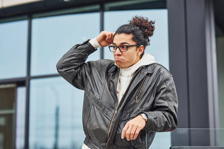 Handsome young man with curly black hair and in glasses is on the street against building.