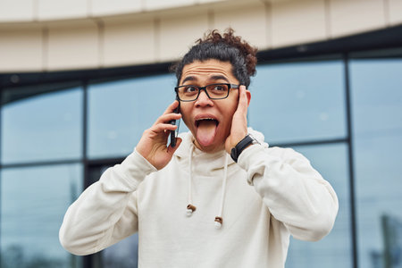 Handsome young man with curly black hair is on the street against building talking by the phone.