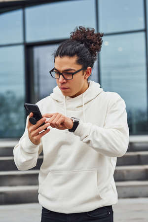 Handsome young man with curly black hair standing on the street with smartphone in hands.の写真素材