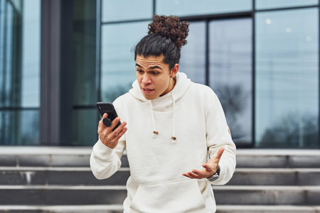 Handsome young man with curly black hair is on the street against building talking by the phone.