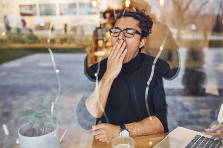 Young guy with black curly hair sitting indoors in cafe. View through the glass.
