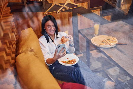 Young brunette sitting indoors with pizza and smartphone in hand.