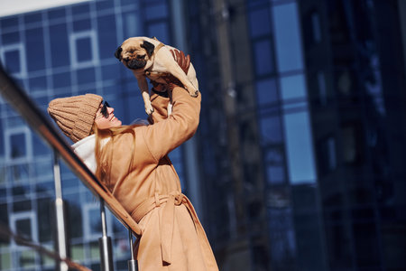 Woman in warm clothes have holding her little pug dog on hands near business building that on background.の写真素材