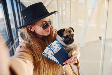 Young female passenger in warm clothes holding tickets and cute dog in hands in airport hall.