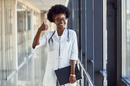Young african american female doctor in white uniform with stethoscope and notepad standing in corridor and showing thumb up.の写真素材