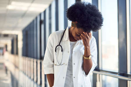 Sad and depressed young african american female doctor in white uniform standing in corridor.の写真素材