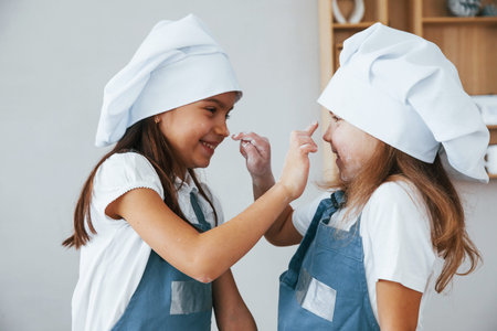 Two little girls in blue chef uniform have fun with flour on the kitchen.