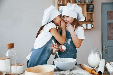 Two little girls in blue chef uniform talking secrets to each other when preparing food on the kitchen.の写真素材