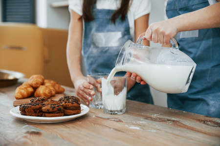 Close up view of two little girls in blue chef uniform that pouring milk into glasses on the kitchen with cookies on table.の写真素材