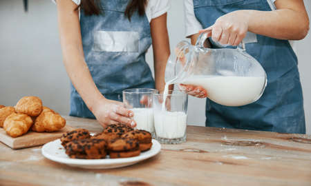 Close up view of two little girls in blue chef uniform that pouring milk into glasses on the kitchen with cookies on table.の写真素材
