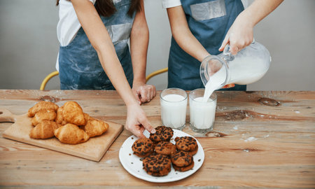 Close up view of two little girls in blue chef uniform that pouring milk into glasses on the kitchen...