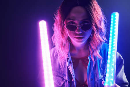 Portrait of young girl in glasses holding light sticks in red and blue neon in studio.の写真素材