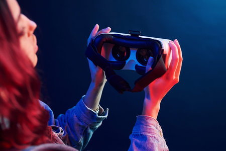 Rear view of girl with curly hair holding virtual reality glasses in red and blue neon in studio.