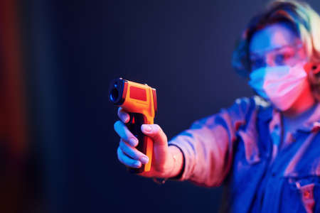 Girl in protective glasses and mask holding infrared thermometer. Neon lights.の写真素材
