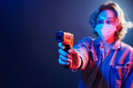 Girl in protective glasses and mask holding infrared thermometer. Neon lights.の写真素材