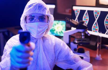 Nurse in mask and white uniform, holding infrared thermometer and sitting in neon lighted laboratory...