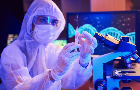 Nurse in mask and white uniform, holding syringe and sitting in neon lighted laboratory with...