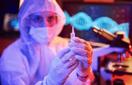 Nurse in mask and white uniform, holding syringe and sitting in neon lighted laboratory with computer and medical equipment searching for Coronavirus vaccine.の写真素材