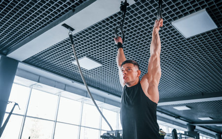 Strong young man in sportive clothes doing exercises on gymnastic rings in the gym.