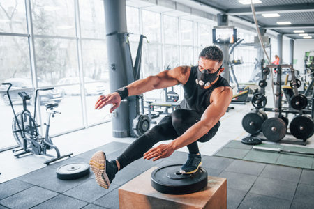 Strong young man in sportive clothes and scary mask doing push ups on one leg in gym.