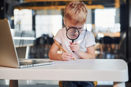 Smart child in casual clothes with laptop on table have fun with magnifying glass.