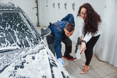 Female customer talking what to do. Male worker in uniform washing new modern car indoors. Conception of service.の写真素材