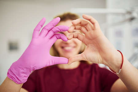 Young female dentist in uniform standing in stomatology office and making heart shaped gesture.の写真素材