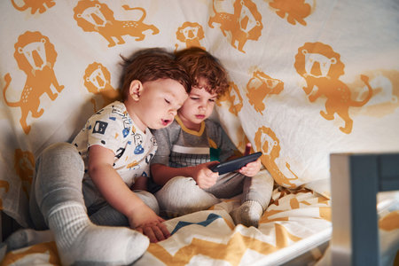 Two little boys resting and have fun indoors under blanket together with smartphone.