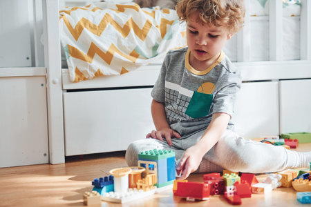 Cute little boy sitting and have fun indoors in the bedroom with plastic construction set.