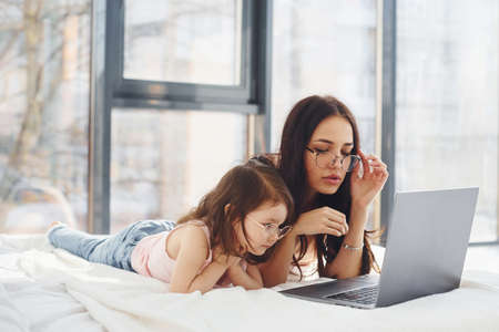 Young mother with her daughter spending weekend together in room and using laptop.の写真素材