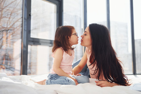 Young mother with her daughter spending weekend together in room and giving a kiss.