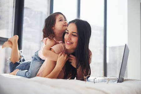 Young mother with her daughter spending weekend together in room and using laptop.の写真素材