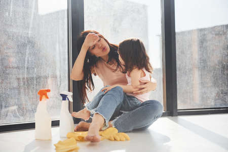Tired woman with her daughter sits on window sill with bottles of cleaning spray and with gloves.の写真素材