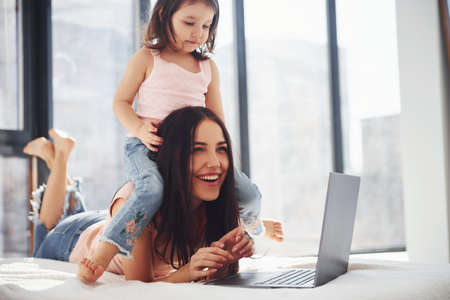 Young mother with her daughter spending weekend together in room and using laptop.の写真素材