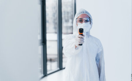 Holds infrared thermometer. Portrait of female doctor scientist in lab coat, defensive eyewear and...
