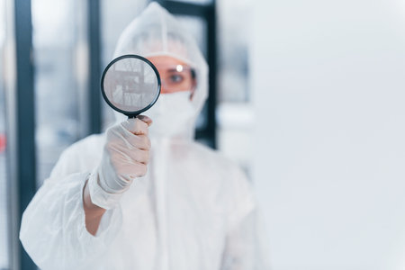 Holds magnifier. Portrait of female doctor scientist in lab coat, defensive eyewear and mask.