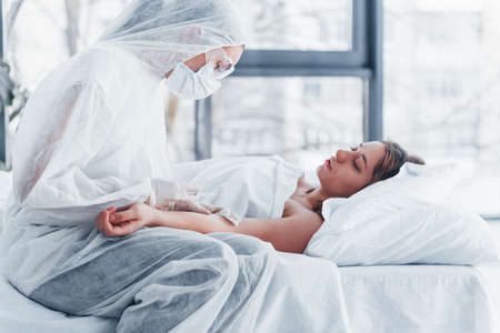 Female doctor in defensive lab coat and protective eyewear with syringe in hand injecting medicine to young girl sick of virus.の写真素材