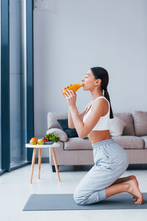 Holds glass with orange juice in hands. Young woman with slim body shape in sportswear have fitness...