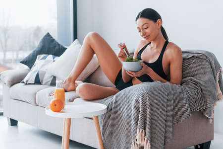 Young woman with slim body shape in sportswear sits on sofa and eats healthy diet food indoors at home.の写真素材