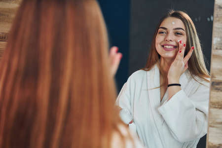 Beautiful young woman standing in bathroom, looking into the mirror and taking care of her face.の写真素材