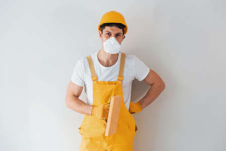 Handyman in yellow uniform and protective mask standing against white wall. House renovation concept.の写真素材