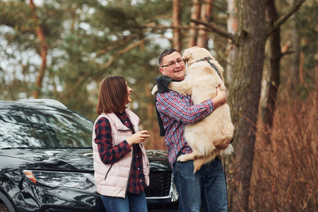 Man with woman have fun with their dog outdoors in forest near modern black car.