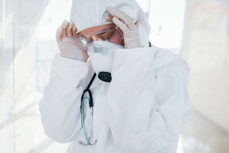 Feels bad and sick. Female doctor scientist in lab coat, defensive eyewear and mask standing indoors.の写真素材