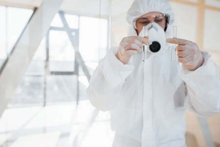 With test tube in hand. Female doctor scientist in lab coat, defensive eyewear and mask standing indoors.の写真素材