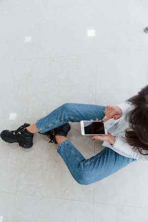Top view of brunette in white shirt with phone in hands that sits indoors in modern airport or hallway at daytime.の写真素材