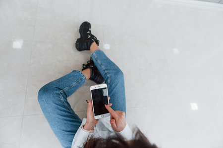 Top view of brunette in white shirt with phone in hands that sits indoors in modern airport or...