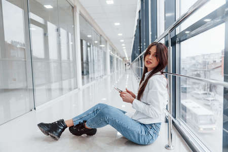 Brunette in white shirt with phone in hands sits indoors in modern airport or hallway at daytime.の写真素材