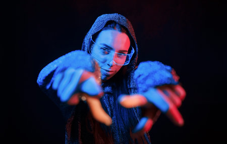 Does gestures by hands. Portrait of young girl that is in eyeglasses in blue neon lighting
