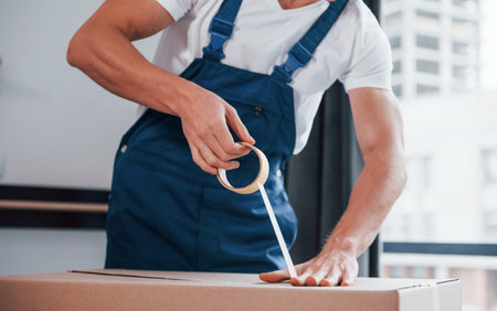Packaging the box. Young male mover in blue uniform works indoors in the roomの写真素材